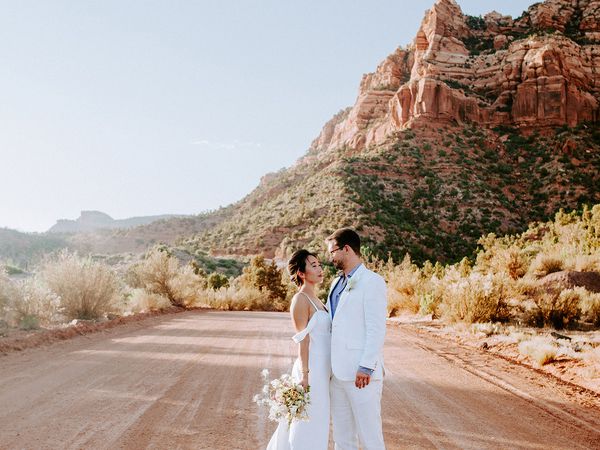 Bride in white wedding dress and groom in white tux standing in the road in a national park on a sunny day, surrounded by canyons.