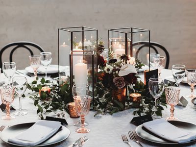 modern square black and glass lantern centerpieces atop greenery, white, and red flowers, at a table set with metallic dining ware.