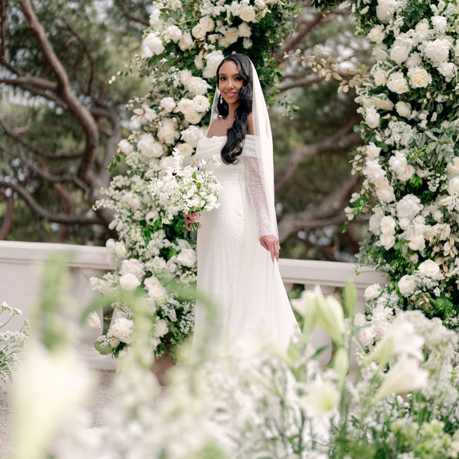 bride posing in front of a white rose altar arch, while holding a lily of the valley wedding bouquet 
