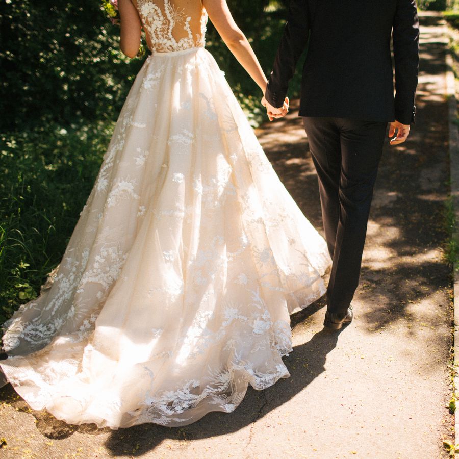 bride in lace wedding dress with see-through back walking with groom on shaded path