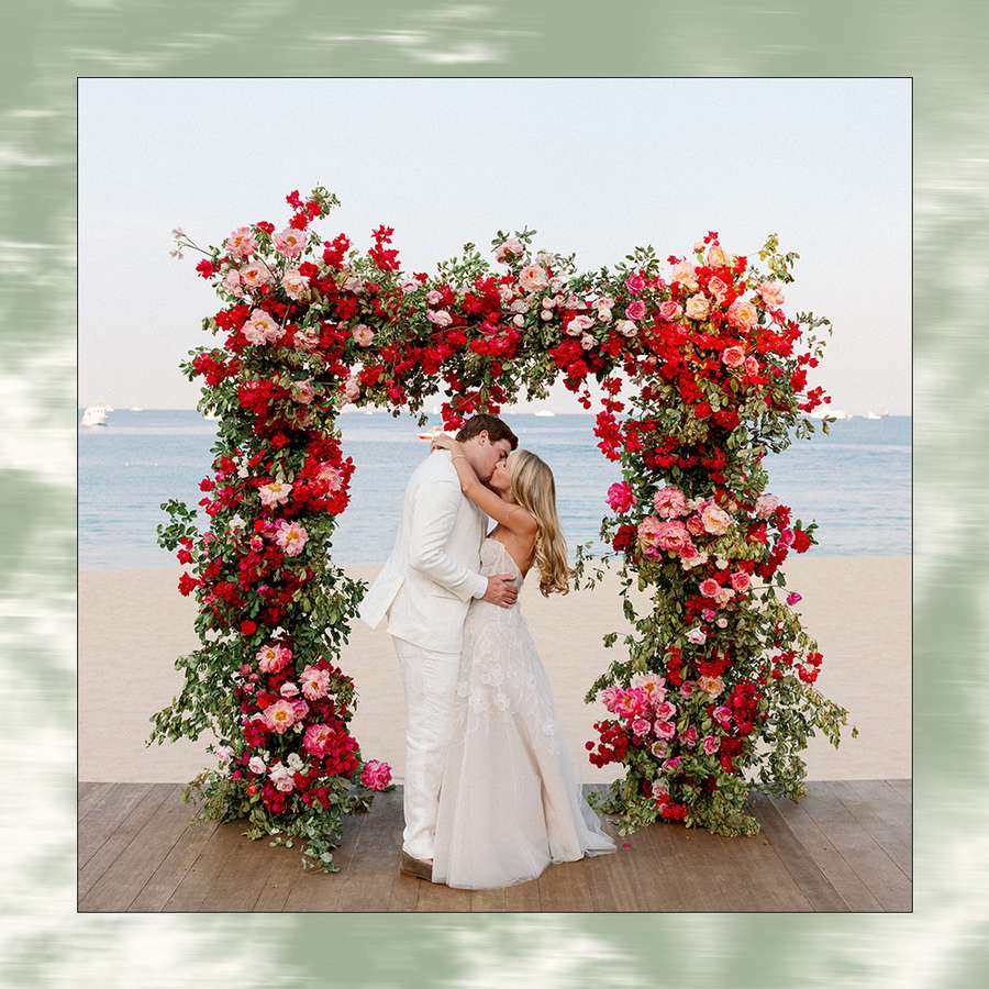 Bride and Groom Kissing Beneath Pink and Red Floral Ceremony Altar at Beachfront Wedding Ceremony