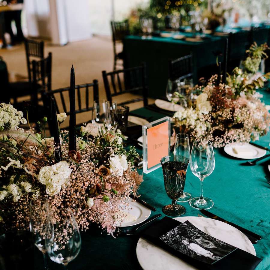 A moody wedding color palette featuring dark teal tablecloths, moody floral bouquets, and glassware.