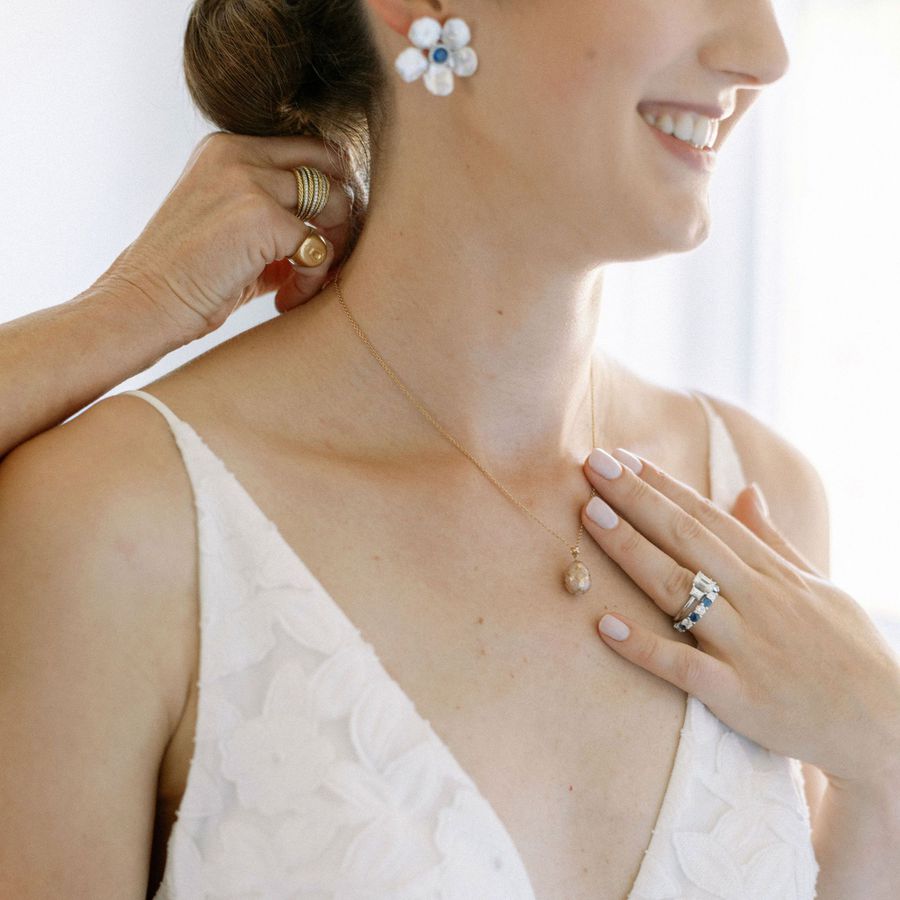 photograph of a smiling bride wearing a deep V-neckline wedding dress, daisy-inspired earrings with a sapphire center stone, a drop pendant necklace, and wedding rings 