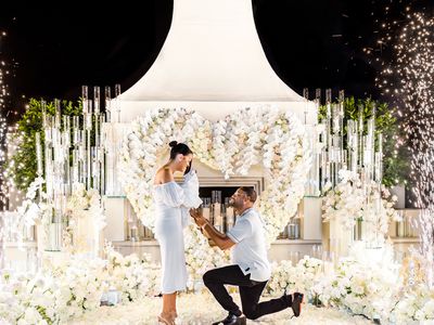 groom on one knee proposing to bride dressed in white and standing in front of fireplace decorate with heart-shaped floral arrangement and candles