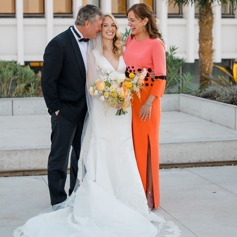 Mother-of-the-bride in coral dress embracing bride and father-of-the-bride