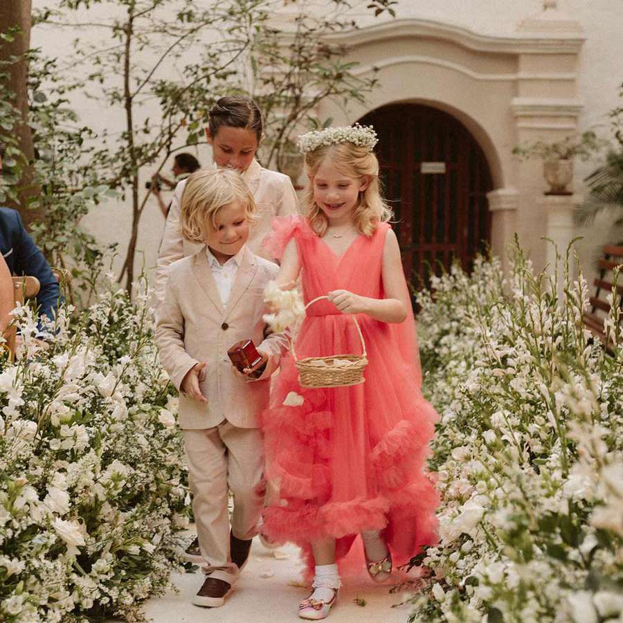 Flower girl in pink dress with flower crown and basket and two ring bearers in tan suits walking down aisle of white florals