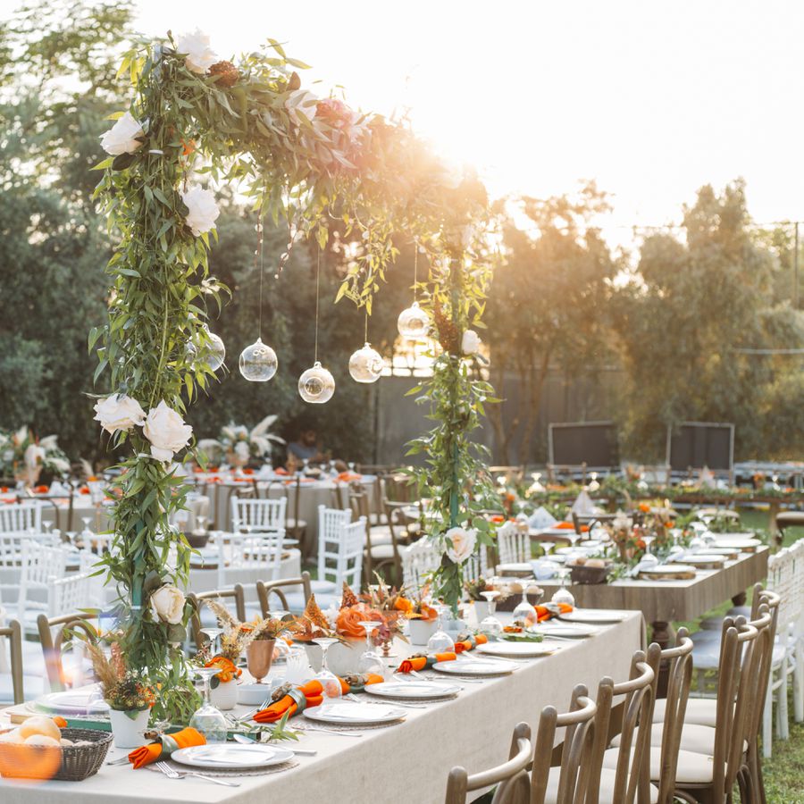 A wedding reception dinner table setup outdoors with white flowers, hanging lights, and greenery.