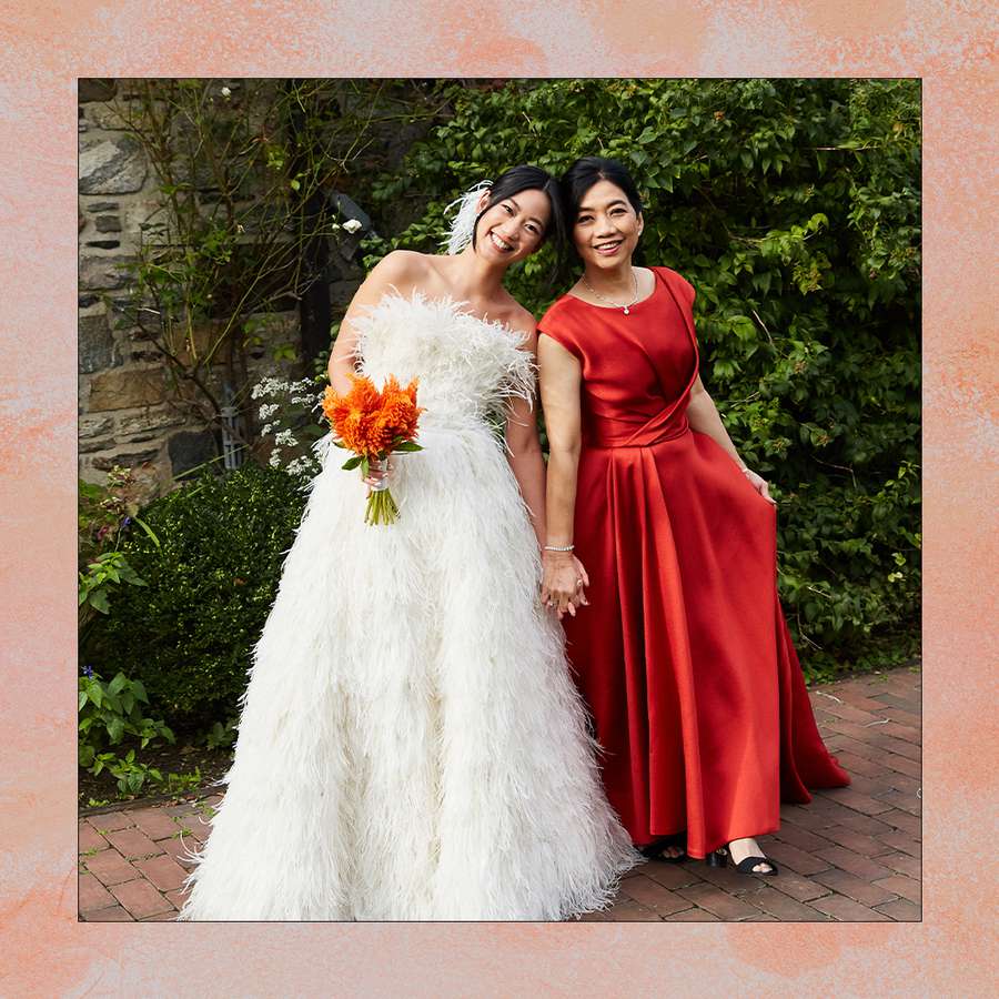 a bride wearing a feather-embellished wedding dress holding hands with her mother who is wearing a red gown