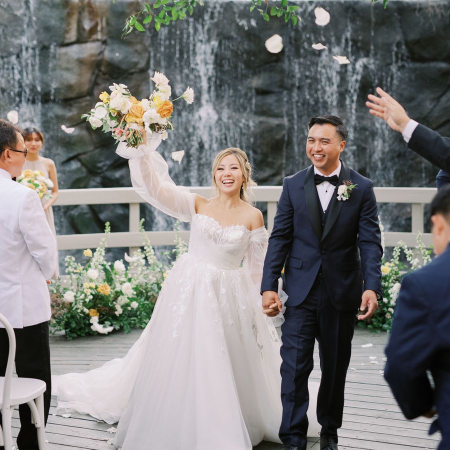 A bride and groom holding hands and smiling during their recessional, while guests toss flower petals at them