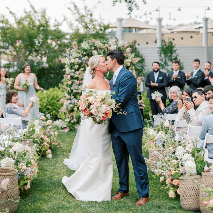 Bride and groom kissing in the middle of their aisle surrounded by wicker baskets and colorful flowers