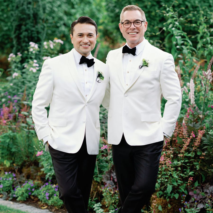 two grooms in white tuxedos with black bow ties and unique boutonniÃ¨res in garden