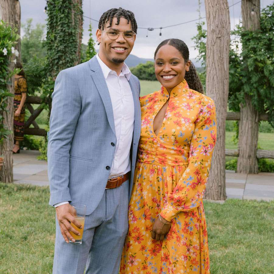 a man wearing a light blue suit and a woman wearing an orange, floral-printed dress to an outdoor wedding