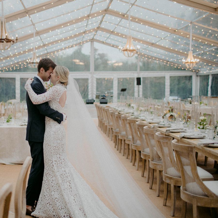Bride and groom embracing, standing together under string lights in clear-top tent wedding venue with long wooden tables.
